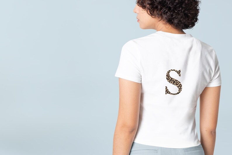 Pode incluir: Camiseta branca com um "S" estampado com estampa de leopardo nas costas. A camiseta de manga curta tem gola redonda. A estampa de leopardo &eacute; uma mistura de manchas marrons e pretas. O fundo &eacute; azul claro.
