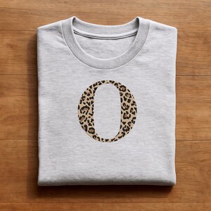 Pode incluir: Camisola cinza claro com gola redonda e mangas compridas. Apresenta uma grande letra "O" bordada com um padr&atilde;o de leopardo em tons de castanho e preto. O design est&aacute; centrado no peito.