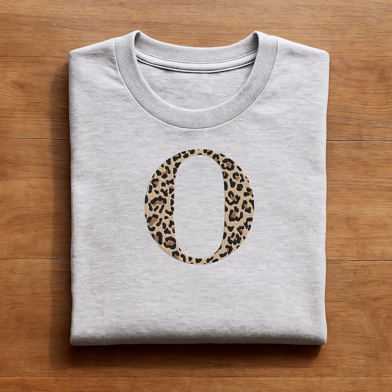 Pode incluir: Camisola cinza claro com gola redonda e mangas compridas. Apresenta uma grande letra "O" bordada com um padr&atilde;o de leopardo em tons de castanho e preto. O design est&aacute; centrado no peito.
