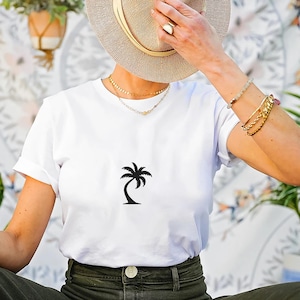 May include: White t-shirt with a black palm tree graphic. The shirt is made of a soft material and is suitable for casual wear. The model is wearing a straw hat and gold jewelry.