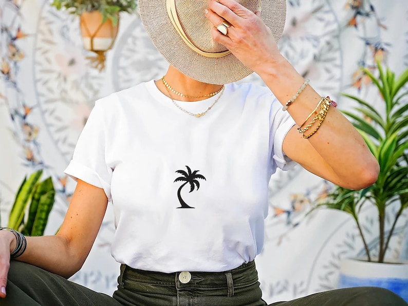 May include: White t-shirt with a black palm tree graphic. The shirt is made of a soft material and is suitable for casual wear. The model is wearing a straw hat and gold jewelry.