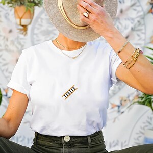 Pu&ograve; includere: T-shirt bianco con una piccola grafica a scala marrone e oro. L'outfit &egrave; abbinato a pantaloni verde oliva e un cappello di paglia. Collane e bracciali dorati.