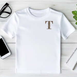 Pode incluir: Camiseta branca com a letra "T" estampada com estampa de leopardo no peito. A camiseta &eacute; exibida em uma superf&iacute;cie de madeira branca, junto com &oacute;culos com arma&ccedil;&atilde;o preta, um smartphone, um bloco de notas, uma caneta e uma pequena planta em vaso.