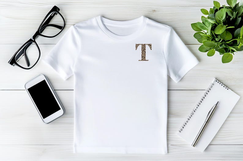 Pode incluir: Camiseta branca com a letra "T" estampada com estampa de leopardo no peito. A camiseta &eacute; exibida em uma superf&iacute;cie de madeira branca, junto com &oacute;culos com arma&ccedil;&atilde;o preta, um smartphone, um bloco de notas, uma caneta e uma pequena planta em vaso.