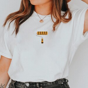 Pu&ograve; includere: T-shirt bianca con grafica a rullo. Il design del rullo presenta strisce gialle e nere sul rullo e un manico giallo e nero. Un capo casual per l'uso quotidiano.