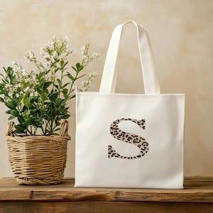 Pode incluir: Uma sacola branca com uma grande letra "S" com estampa de leopardo na frente. A sacola tem duas al&ccedil;as brancas e est&aacute; sobre uma superf&iacute;cie de madeira. &Agrave; esquerda da sacola h&aacute; uma cesta de vime com uma planta verde e flores brancas.