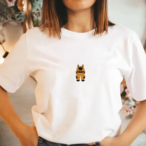 Pu&ograve; includere: T-shirt bianca con un piccolo personaggio dei cartoni animati ricamato in marrone, nero e giallo. Il personaggio indossa un'uniforme o un abito. La maglietta &egrave; un classico girocollo.