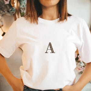 Pode incluir: Uma camiseta branca com a letra "A" estampada com estampa de leopardo. A camiseta tem mangas curtas e gola redonda. A pessoa est&aacute; usando jeans azul.