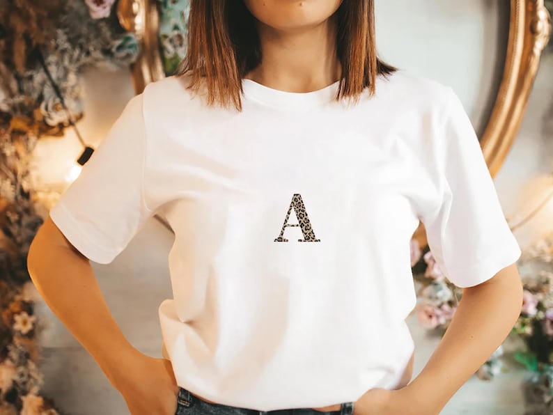 Pode incluir: Uma camiseta branca com a letra "A" estampada com estampa de leopardo. A camiseta tem mangas curtas e gola redonda. A pessoa est&aacute; usando jeans azul.