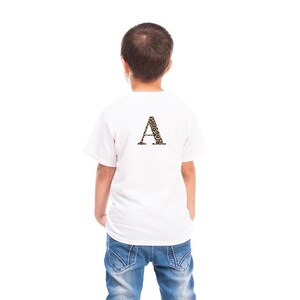 Pode incluir: Uma camiseta branca com uma grande letra "A" com estampa de leopardo nas costas. A camiseta &eacute; usada por uma crian&ccedil;a, que est&aacute; de costas para a c&acirc;mera. A crian&ccedil;a est&aacute; usando jeans azuis.
