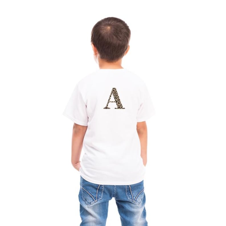 Pode incluir: Uma camiseta branca com uma grande letra "A" com estampa de leopardo nas costas. A camiseta &eacute; usada por uma crian&ccedil;a, que est&aacute; de costas para a c&acirc;mera. A crian&ccedil;a est&aacute; usando jeans azuis.