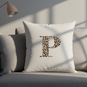 Pode incluir: Uma almofada quadrada branca com a letra "P" num design de estampa de leopardo. A almofada est&aacute; num sof&aacute; cinzento. Uma l&acirc;mpada e uma planta est&atilde;o no fundo.