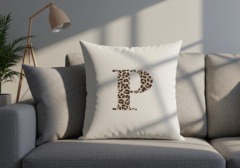 Pode incluir: Uma almofada quadrada branca com a letra "P" num design de estampa de leopardo. A almofada est&aacute; num sof&aacute; cinzento. Uma l&acirc;mpada e uma planta est&atilde;o no fundo.