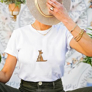 May include: White t-shirt featuring a cartoon kangaroo in shades of brown. The shirt is made of a soft material and is suitable for casual wear. The kangaroo is depicted in a simple, minimalist style.