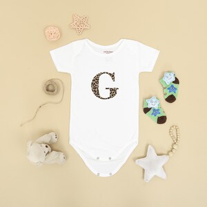 Pode incluir: Body de beb&eacute; branco com um "G" com estampa de leopardo. A imagem inclui meias com estrelas, uma chocalho em forma de estrela e um ursinho de pel&uacute;cia, sobre um fundo bege.