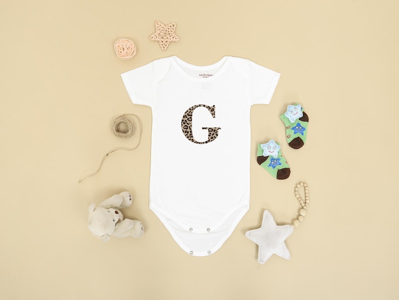 Pode incluir: Body de beb&eacute; branco com um "G" com estampa de leopardo. A imagem inclui meias com estrelas, uma chocalho em forma de estrela e um ursinho de pel&uacute;cia, sobre um fundo bege.