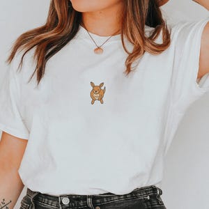 May include: White t-shirt featuring a small, cartoon-style kangaroo graphic in shades of brown. The kangaroo is centered on the chest. The shirt is made of a soft, comfortable material. A casual, everyday wear tee.