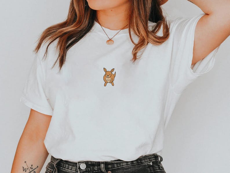 May include: White t-shirt featuring a small, cartoon-style kangaroo graphic in shades of brown. The kangaroo is centered on the chest. The shirt is made of a soft, comfortable material. A casual, everyday wear tee.