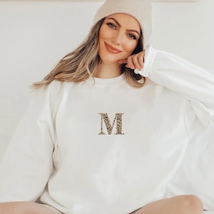 Pode incluir: Camisola branca de manga comprida com um "M" estampado em leopardo bordado na frente. A camisola &eacute; feita de um material macio e tem gola redonda. A modelo usa um gorro bege.