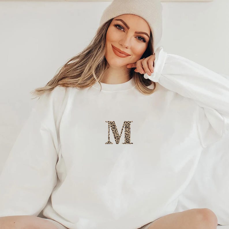 Pode incluir: Camisola branca de manga comprida com um "M" estampado em leopardo bordado na frente. A camisola &eacute; feita de um material macio e tem gola redonda. A modelo usa um gorro bege.