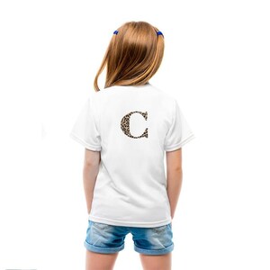 Pode incluir: Uma camiseta branca com a letra "C" estampada em leopardo nas costas. A camisa &eacute; usada com shorts jeans. A pessoa tem cabelo castanho e usa la&ccedil;os de cabelo azuis.