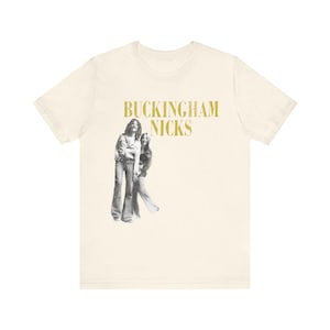 Puede incluir: Camiseta color crema con una imagen en tonos sepia de dos personas y el texto "BUCKINGHAM NICKS" en letras doradas. La camiseta gráfica presenta una estética vintage, ideal para los amantes de la música.