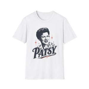 Pode incluir: Uma t-shirt branca de manga curta com um retrato a preto e branco de uma mulher de cabelo escuro. Abaixo do retrato, o nome PATSY está impresso numa fonte azul escura com um sublinhado vermelho. Pequenos detalhes de estrelas vermelhas rodeiam o retrato.
