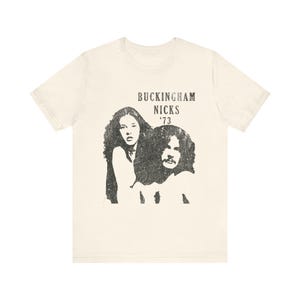 May include: Off-white t-shirt with a black and white graphic of two people and the text "BUCKINGHAM NICKS '73". The graphic has a vintage, distressed appearance. The shirt is made of a soft material.