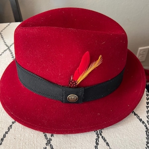 Peut inclure: Un chapeau fedora rouge avec un ruban noir et une plume d&eacute;corative. Le chapeau a une couronne ronde et un bord. Un petit embl&egrave;me dor&eacute; est sur le ruban noir. Le chapeau est fait d'une mati&egrave;re semblable &agrave; du feutre.
