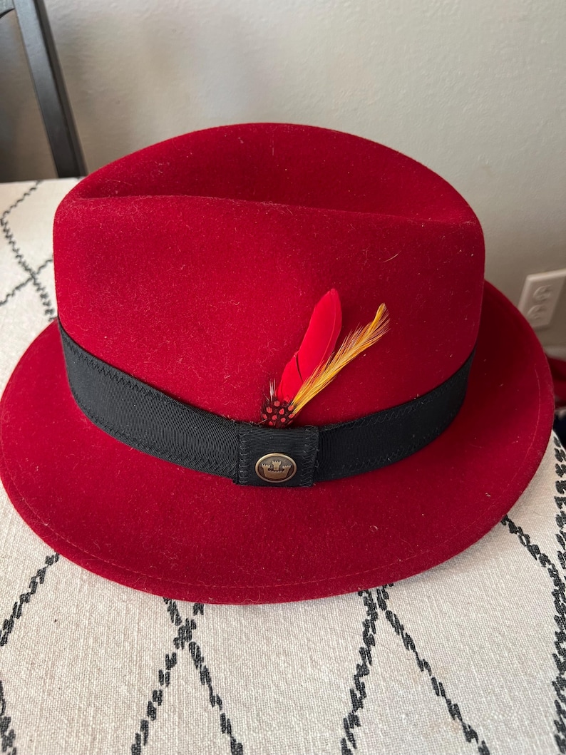 Peut inclure: Un chapeau fedora rouge avec un ruban noir et une plume d&eacute;corative. Le chapeau a une couronne ronde et un bord. Un petit embl&egrave;me dor&eacute; est sur le ruban noir. Le chapeau est fait d'une mati&egrave;re semblable &agrave; du feutre.