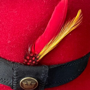 Peut inclure: Un chapeau fedora rouge vif avec un ruban en gros-grain noir. Le chapeau est orn&eacute; de deux plumes, une rouge et une jaune. Un embl&egrave;me m&eacute;tallique dor&eacute; avec un motif de couronne est fix&eacute; au ruban.