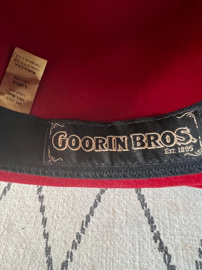 Peut inclure: Gros plan sur un chapeau en laine rouge avec une bande int&eacute;rieure noire. Le chapeau porte une &eacute;tiquette avec le texte "GOORIN BROS. EST. 1895". Le chapeau est &eacute;tiquet&eacute; "LARGE" et fabriqu&eacute; aux &Eacute;tats-Unis.