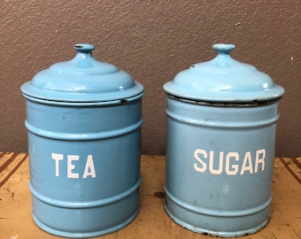 May include: Two vintage blue metal canisters with white lids and white lettering. The canister on the left says "TEA" and the canister on the right says "SUGAR".