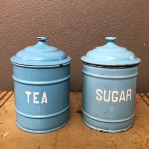 May include: Two vintage blue metal canisters with white lids and white lettering. The canister on the left says "TEA" and the canister on the right says "SUGAR".