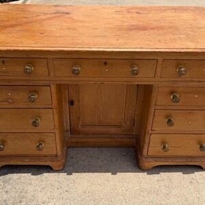 May include: A rectangular wooden desk with a natural light brown finish, featuring a flat top surface, three top drawers, and two columns of three drawers each flanking a central cabinet door. Each drawer and the cabinet door have round brass knobs.