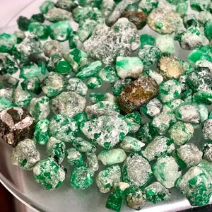 May include: A collection of raw emerald gemstones in various shapes and sizes. The stones display a range of green hues, from vibrant emerald to lighter shades, with some showing a crystalline structure. Some stones have a coating of white or gray material.
