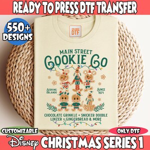 May include: Cream-colored t-shirt with a Christmas-themed design. The shirt features the text "Main Street Gookie Co" and gingerbread characters. Additional text includes "Serving Orlando" and "Since 1971". The shirt is part of the "Disney Christmas Series 1".