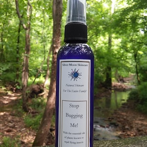 Puede incluir: Una botella de spray azul con boquilla negra y tapa transparente. La etiqueta dice "Silver Moon Skincare" y "Stop Bugging Me!" con el texto "4oz". El fondo es una vista borrosa de un bosque y un arroyo.