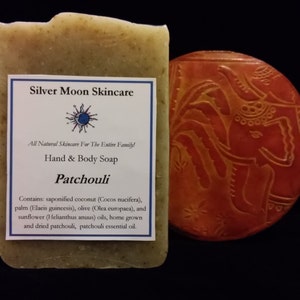 Peut inclure: Un savon naturel pour les mains et le corps avec une étiquette indiquant "Silver Moon Skincare" et "Patchouli". Le savon est sur une surface brune à côté d'un dessous-de-verre rond, orange et décoratif avec un motif en relief.