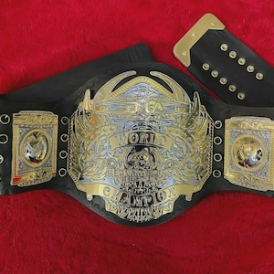 Puede incluir: Un cinturón de campeonato de lucha libre con una correa de cuero negro y detalles metálicos dorados y plateados. El cinturón presenta el texto "NWA World Heavyweight Champion" y diseños intrincados.