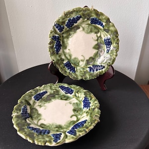 May include: Two ceramic plates with a grape design. The plates have a white center, a green border with grape leaves, and clusters of blue grapes. The plates are on a dark surface.