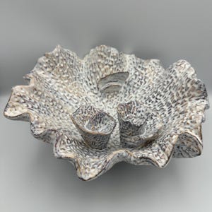 May include: A ceramic bowl with a ruffled, wavy edge and a textured, porous surface. The exterior and interior feature a mottled white and brown finish. Two small, cup-like structures are attached to the interior base of the bowl.