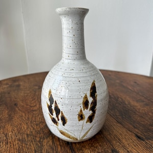 May include: A speckled white ceramic vase with a narrow neck and rounded body. The vase features brown floral designs and is approximately 8 inches tall. The vase is a decorative item for the home.