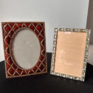 May include: Two rectangular picture frames. The left frame features a deep red enamel finish with a gold-toned lattice pattern and small amber-colored crystals. The right frame has a silver-toned border set with clear, rectangular-cut crystals.