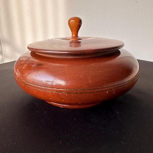 May include: A round, lidded wooden container with a rich, reddish-brown finish. The lid features a small, rounded handle. The container has a smooth, polished surface, suggesting it is a decorative or storage item.