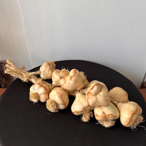 May include: A decorative garlic braid featuring multiple garlic bulbs with tan and brown tones. The bulbs are tied together with a natural fiber rope. The garlic braid is displayed on a black surface.