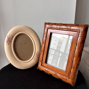 May include: An oval cream-colored picture frame with floral accents on the sides and a rectangular brown picture frame with a woven-look border. Both frames are empty and feature a smooth finish, providing a clear view of the inner display area.