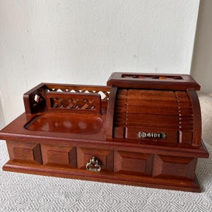 May include: A brown wooden desk organizer featuring a rectangular base with a single drawer, a central open tray, and a raised section on the right side with a curved, slatted roll-top lid. The front of the drawer has a small metal pull handle.
