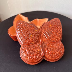 May include: A ceramic butterfly-shaped dish with a lid. The dish is a warm reddish-brown color with intricate embossed details. The lid is open, revealing the interior of the dish, which is a lighter orange hue.