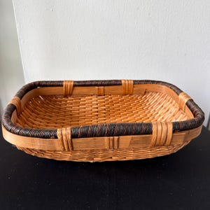 May include: A rectangular woven basket with a light brown base and a dark brown border. The basket is made of natural materials and has a simple, rustic design. It is empty and sits on a black surface.